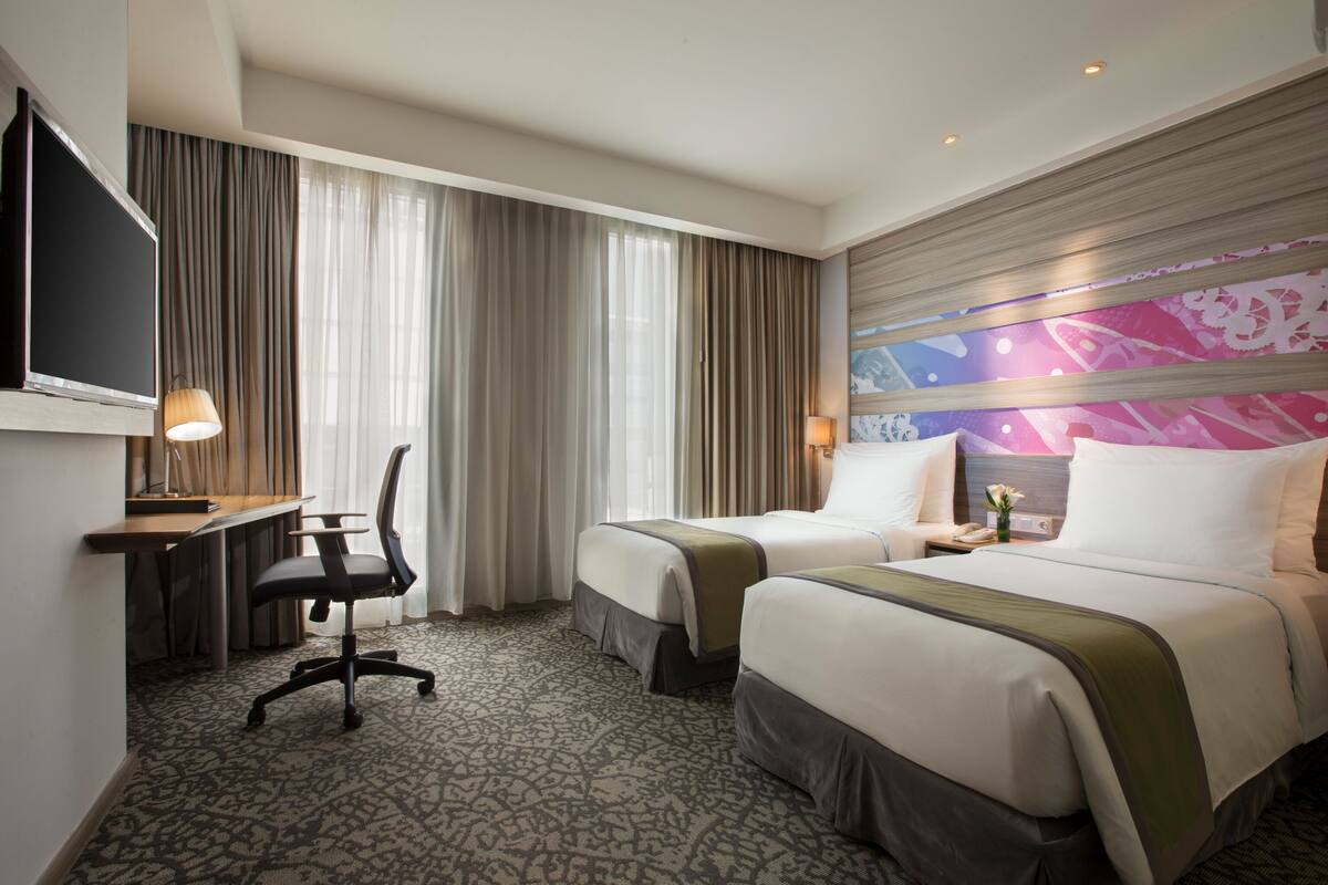 Ein Hotelzimmer mit zwei Betten, einem Schreibtisch mit Stuhl, einem Fernseher und einem großen Fenster mit Vorhängen.