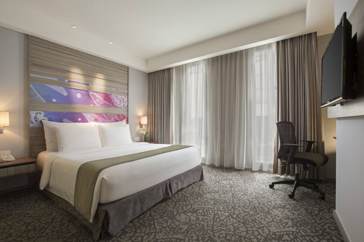 Ein Hotelzimmer mit einem großen Bett, einem Schreibtisch mit Stuhl, einem Flachbildfernseher und einem großen Fenster mit Vorhängen.