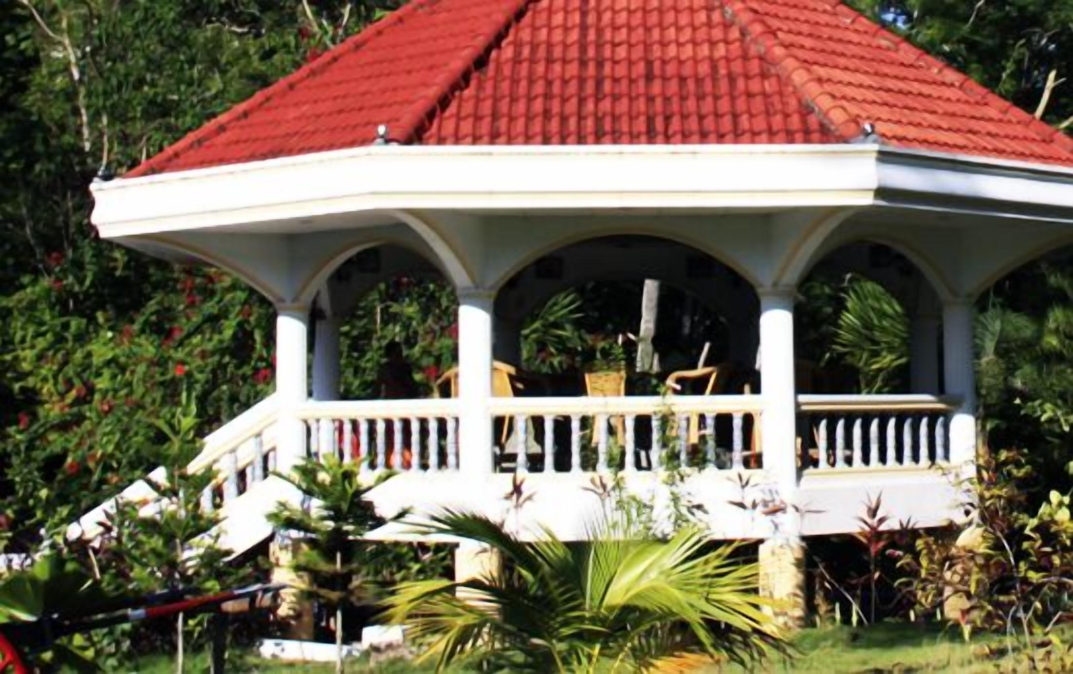 A white gazebo with a red roof, surrounded by lush greenery.