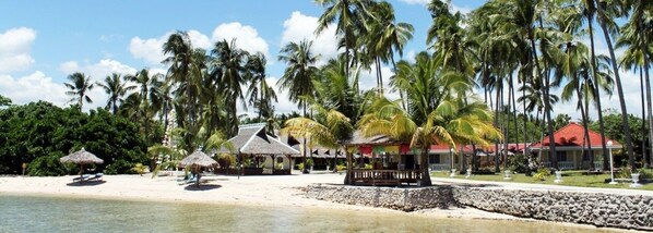 A beach resort with palm trees, thatched-roof huts, and a red-roofed building.