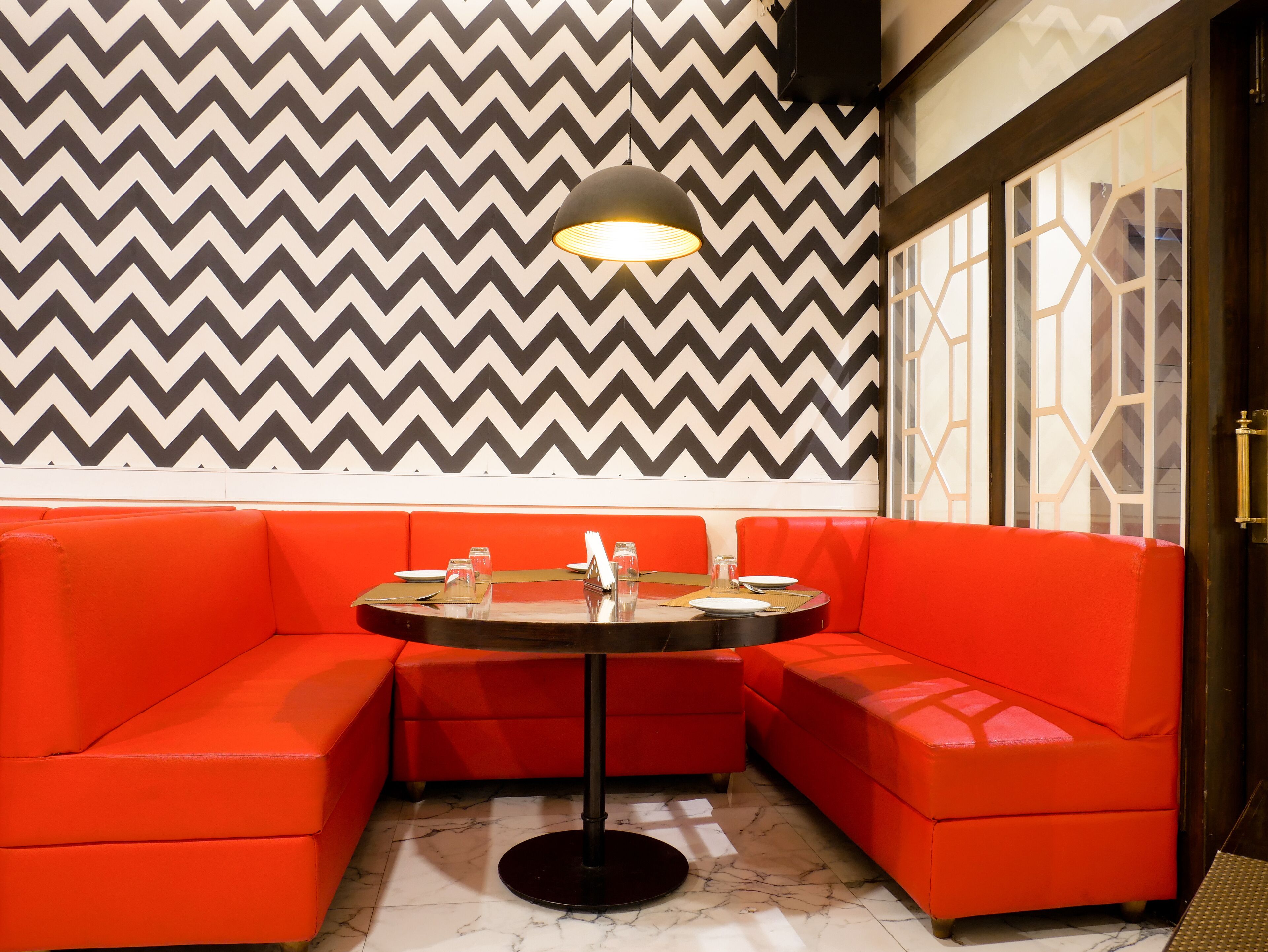 A corner seating area with a round table, red cushioned benches, a black floor lamp, and a chevron-patterned wall.
