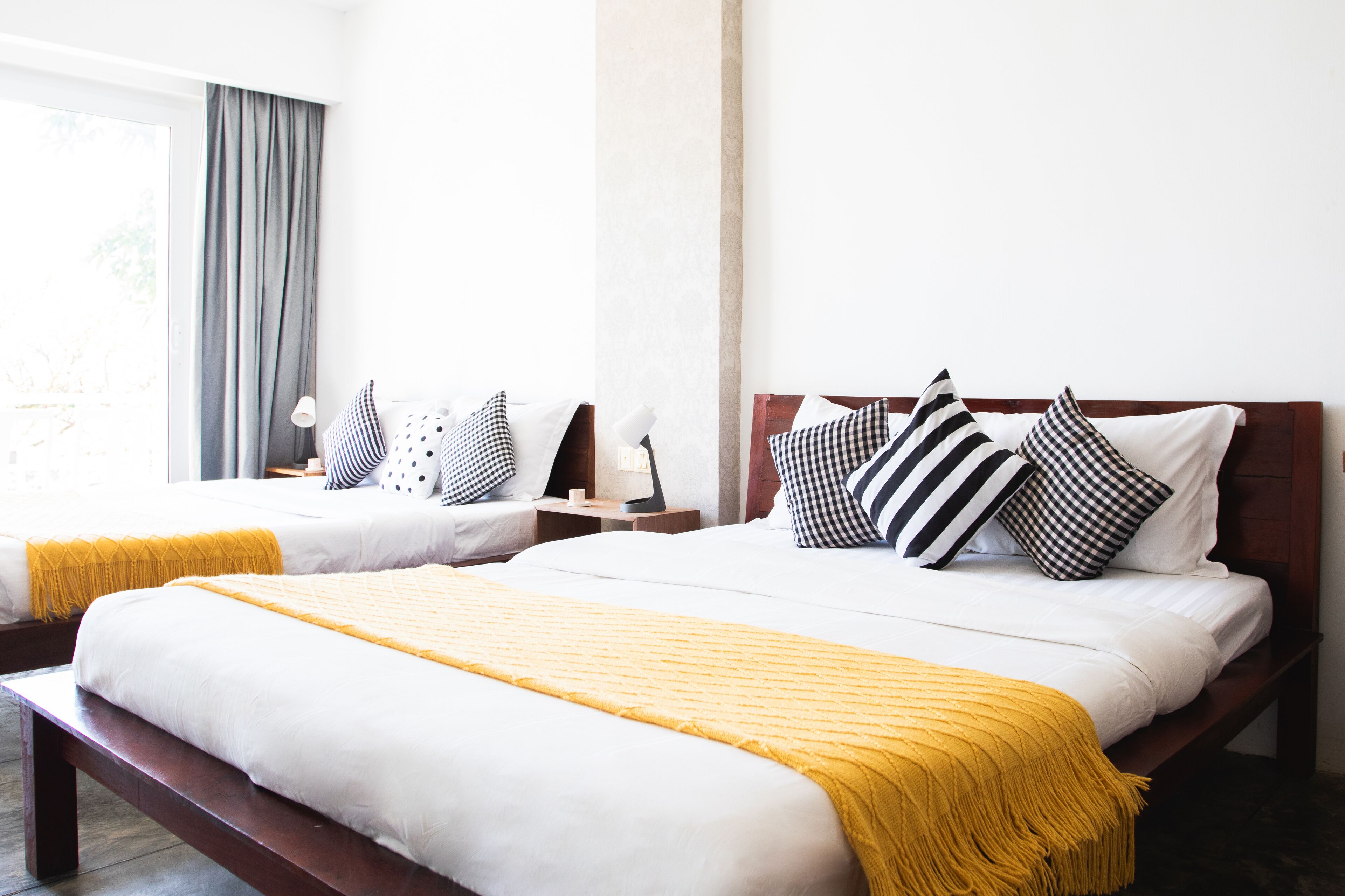 Habitación de hotel con dos camas, cada una con ropa de cama blanca y mantas de color amarillo. Variedades de almohadas con diseños blanco y negro. La habitación tiene un ventanal grande con cortinas grises y una mesita de noche de madera con una lámpara.