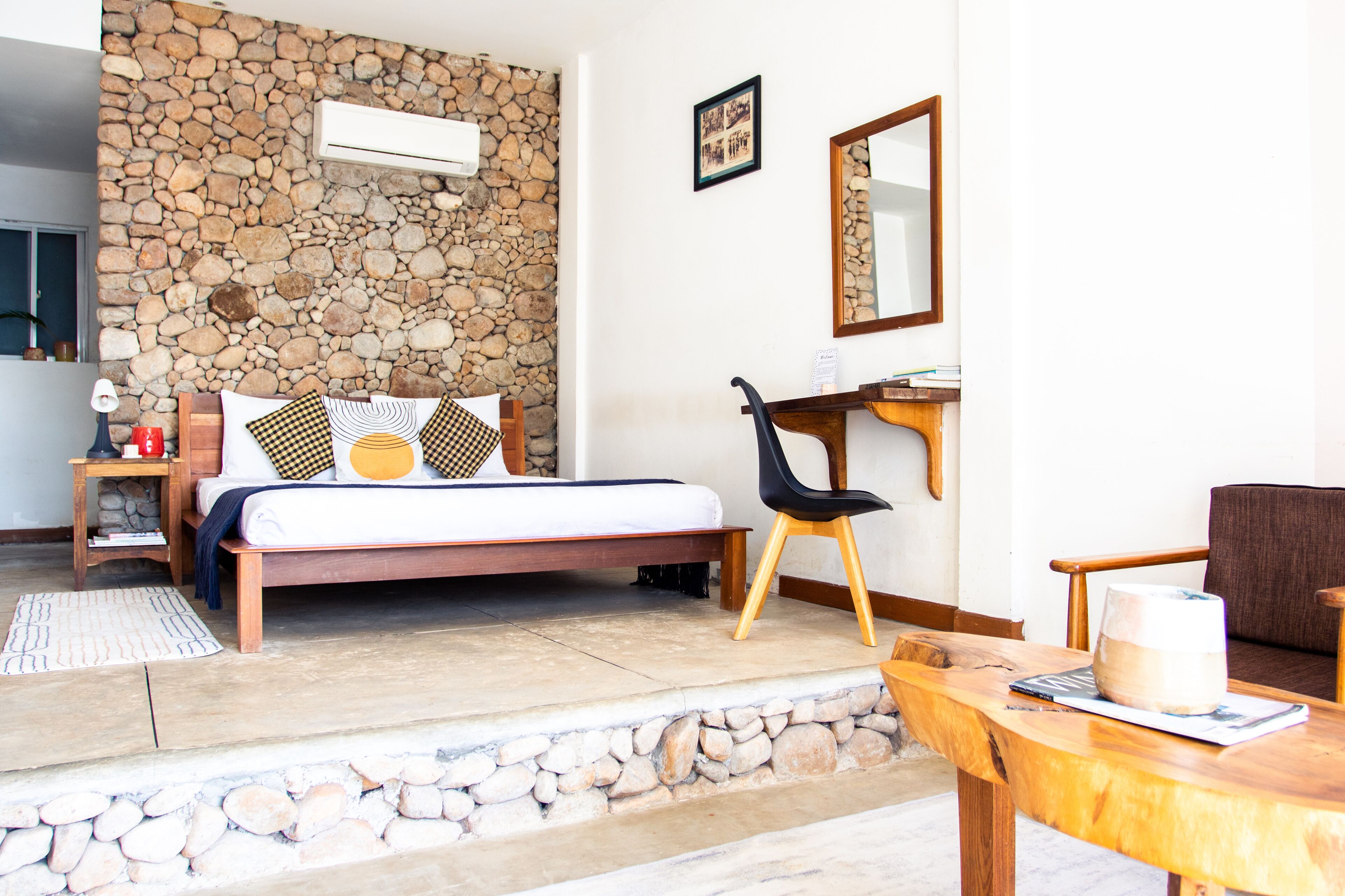 Un dormitorio con pared de piedra, una cama con almohadas estampadas, una mesita de noche de madera, una silla, un escritorio y un espejo.