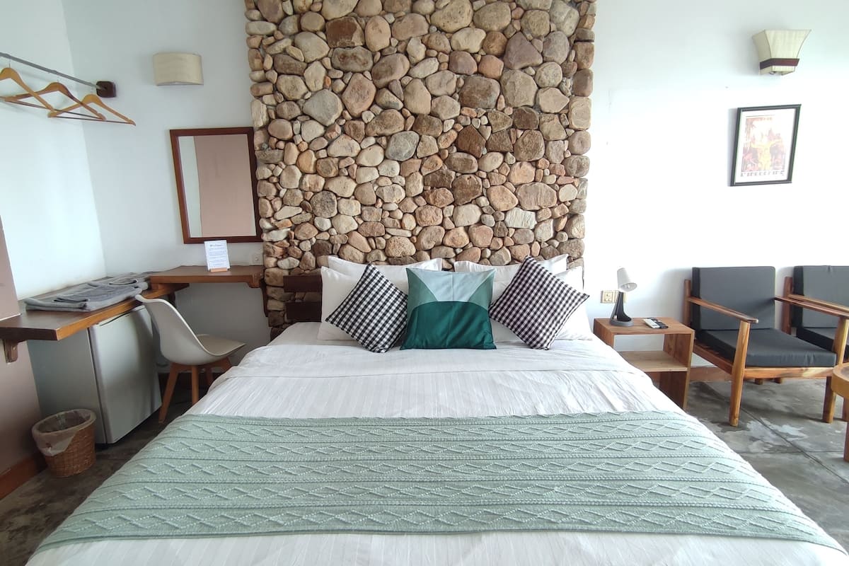 A hotel room with a stone wall, a bed with a green blanket, a desk with a chair, a TV, and a sofa.