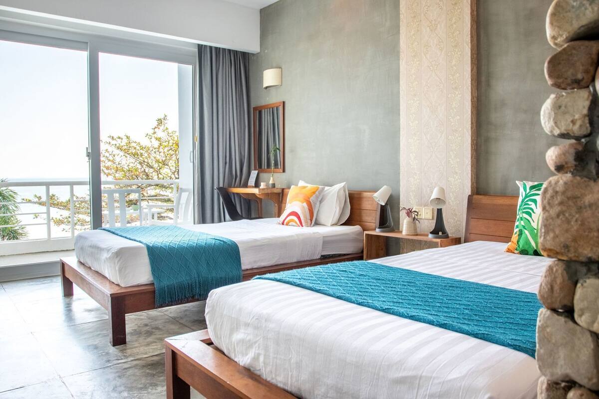 A hotel room with two beds, a balcony, a desk, and a view of the outdoors.