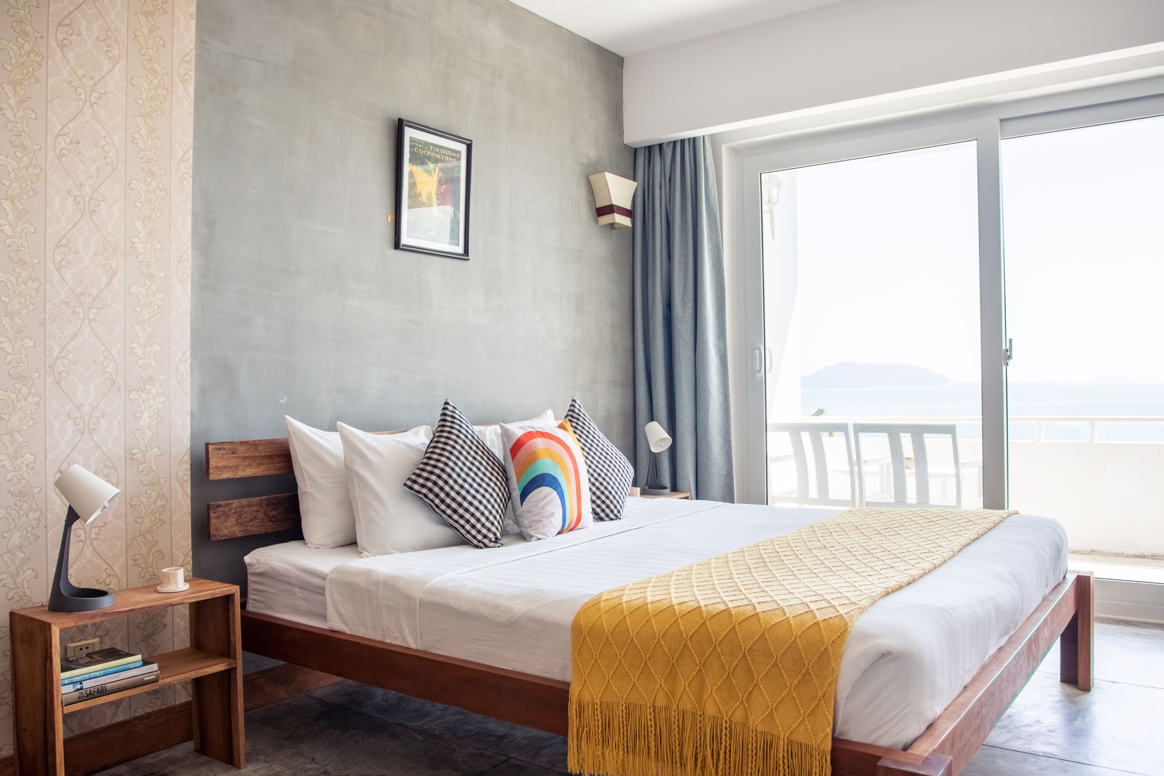 Quarto de hotel moderno com uma cama grande, uma mesinha de cabeceira de madeira, um criado-mudo, uma luminária e vista para o mar através de uma porta de vidro deslizante.
