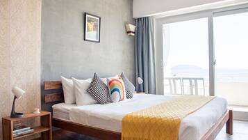 Quarto de hotel moderno com uma cama grande, uma mesinha de cabeceira de madeira, um criado-mudo, uma luminária e vista para o mar através de uma porta de vidro deslizante.