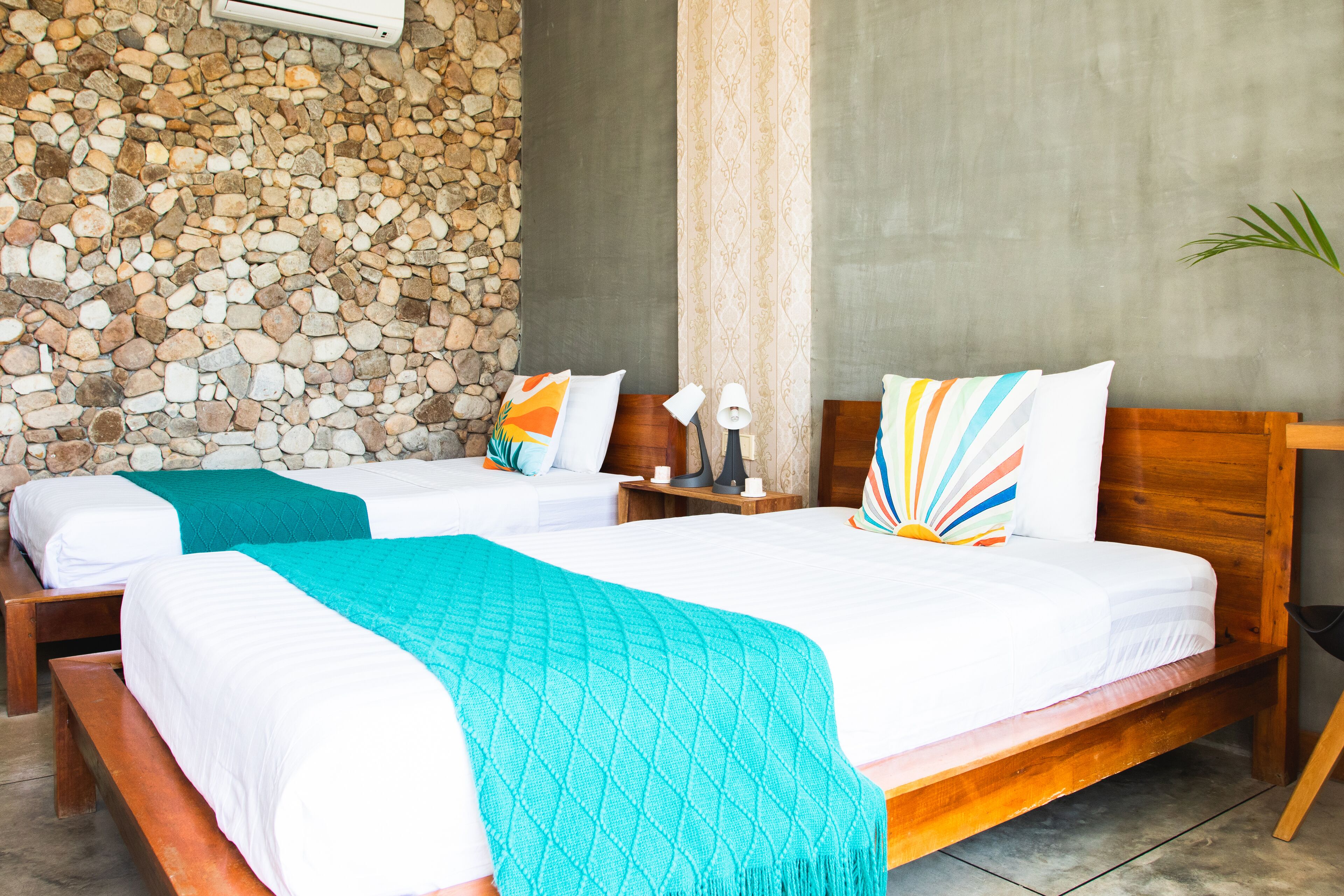 A hotel room with two beds, a stone wall, a wooden bench, and a nightstand with a lamp.