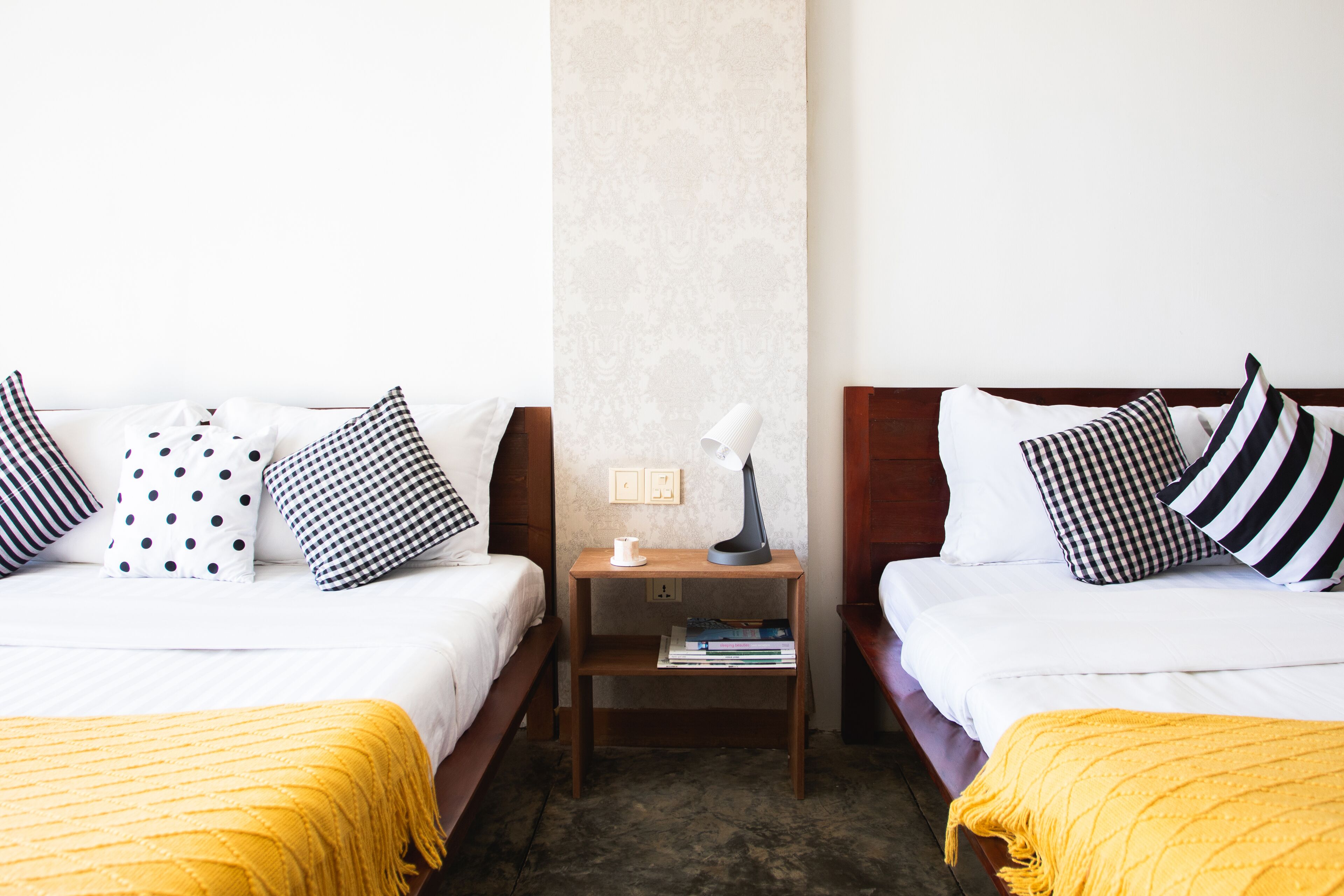 Ein Hotelzimmer mit zwei Betten, jedes mit einer gelben Tagesdecke, und einem Nachttisch dazwischen mit einer Lampe und Büchern.