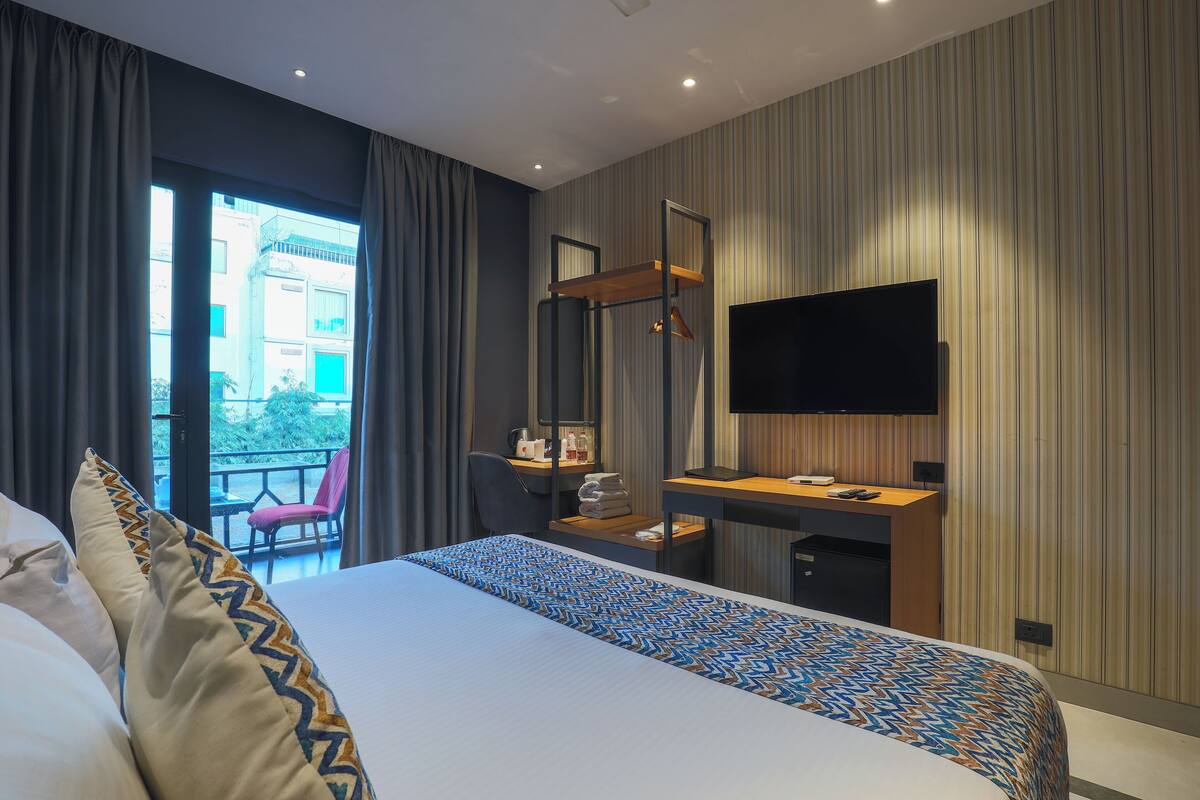 A hotel room with a bed, a desk, a chair, a TV, and a balcony with a view of buildings.