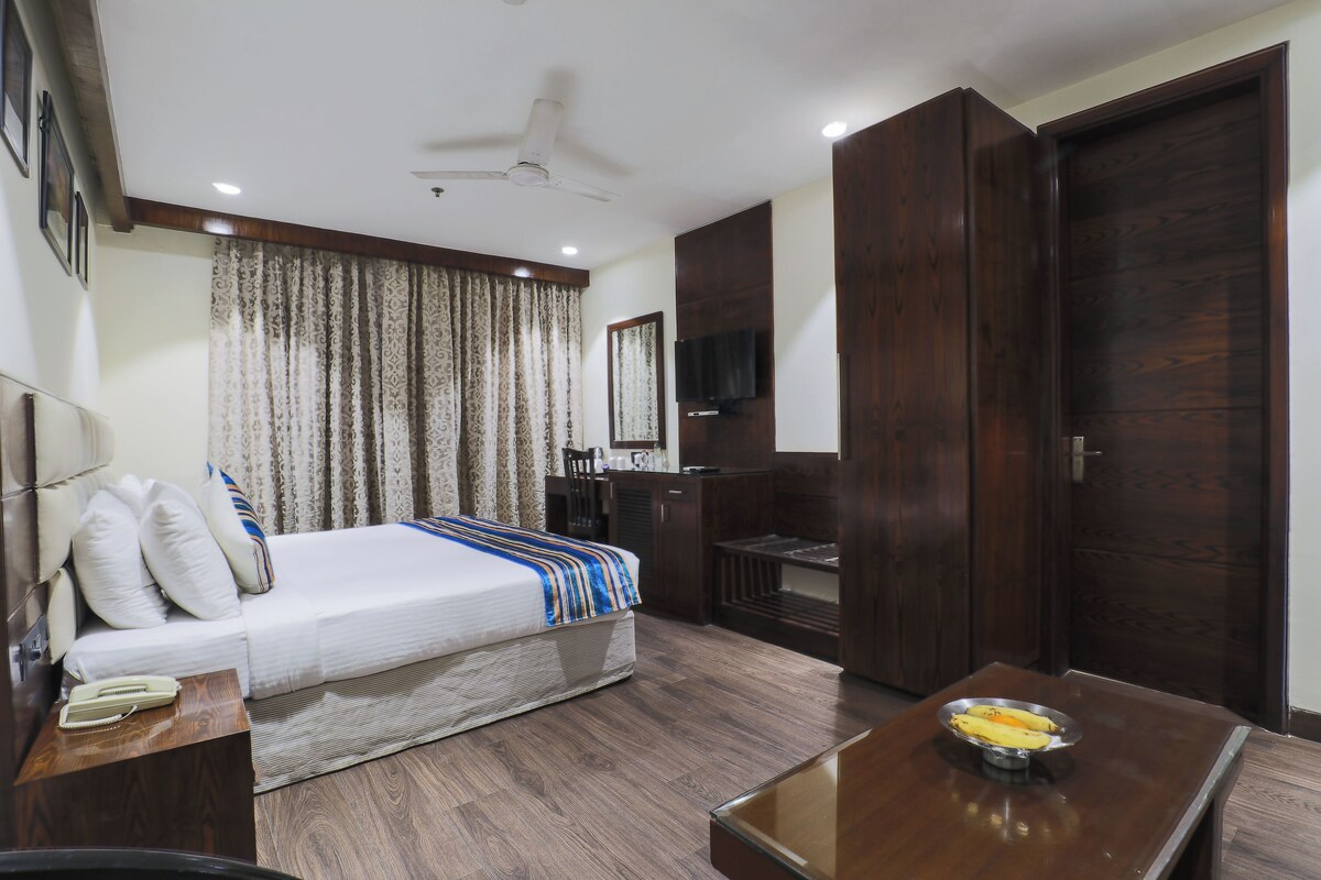 A hotel room with a bed, a TV, a wooden wardrobe, a desk with a bowl of fruit, and a ceiling fan.