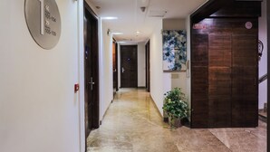 A hotel corridor with a fire exit sign, a plant, and a door with a fire alarm. - Hotel BlueStone - Nehru Place (New Delhi)