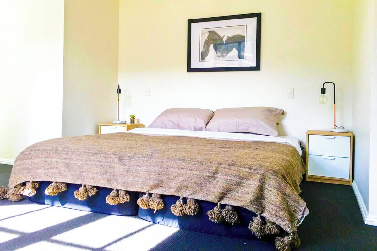 A bedroom with a bed, two bedside tables, a nightstand, a wall-mounted picture, and a window with sunlight.