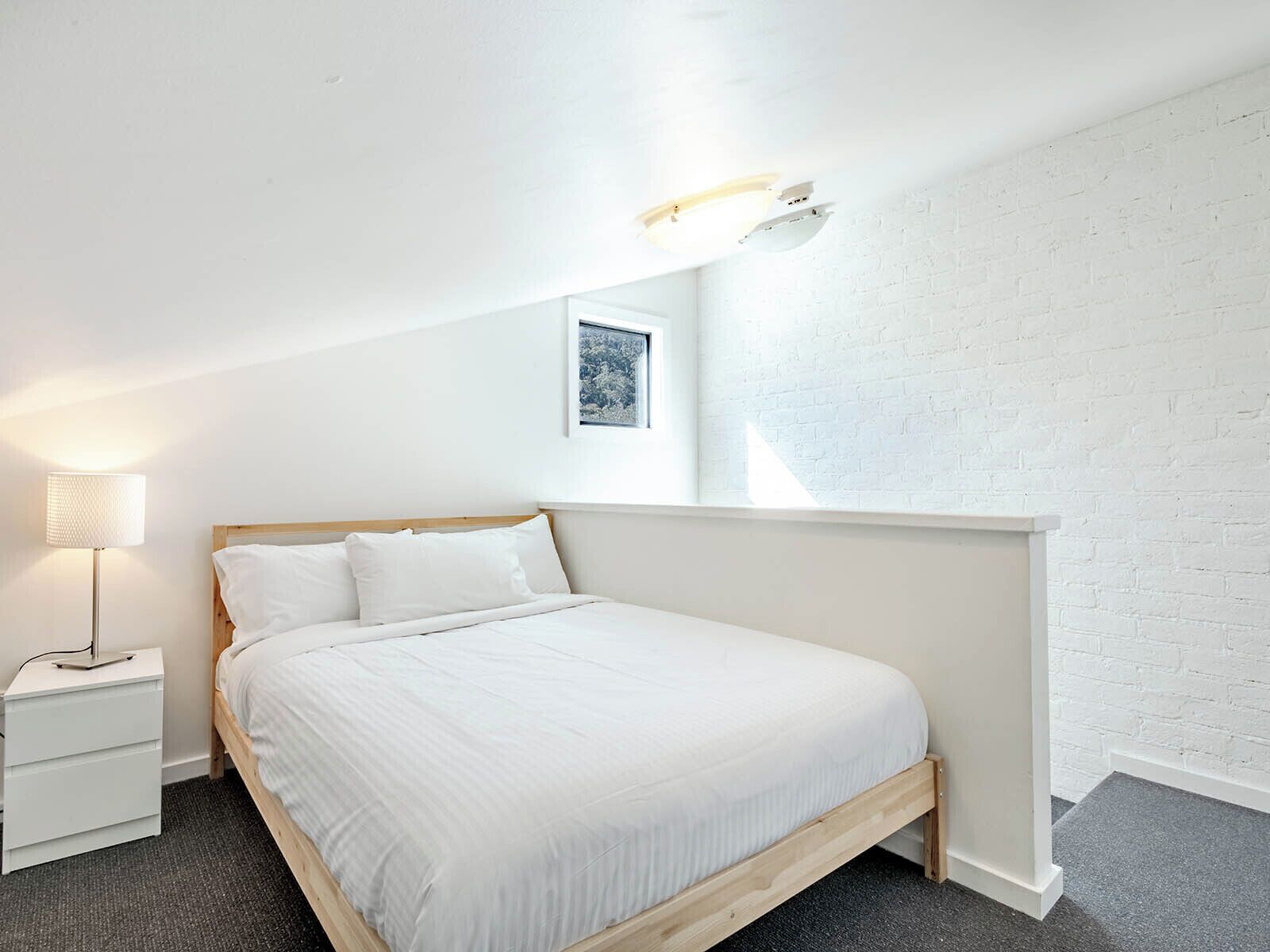 A minimalist bedroom with a wooden bed, white bedding, a nightstand with a lamp, and a white brick wall.