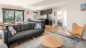 Ein modernes Wohnzimmer mit grauem Sofa, Holztisch und einem Essbereich mit Stühlen. Im Hintergrund ist eine Küche mit dunklen Schränken und einem Edelstahlkühlschrank zu sehen.