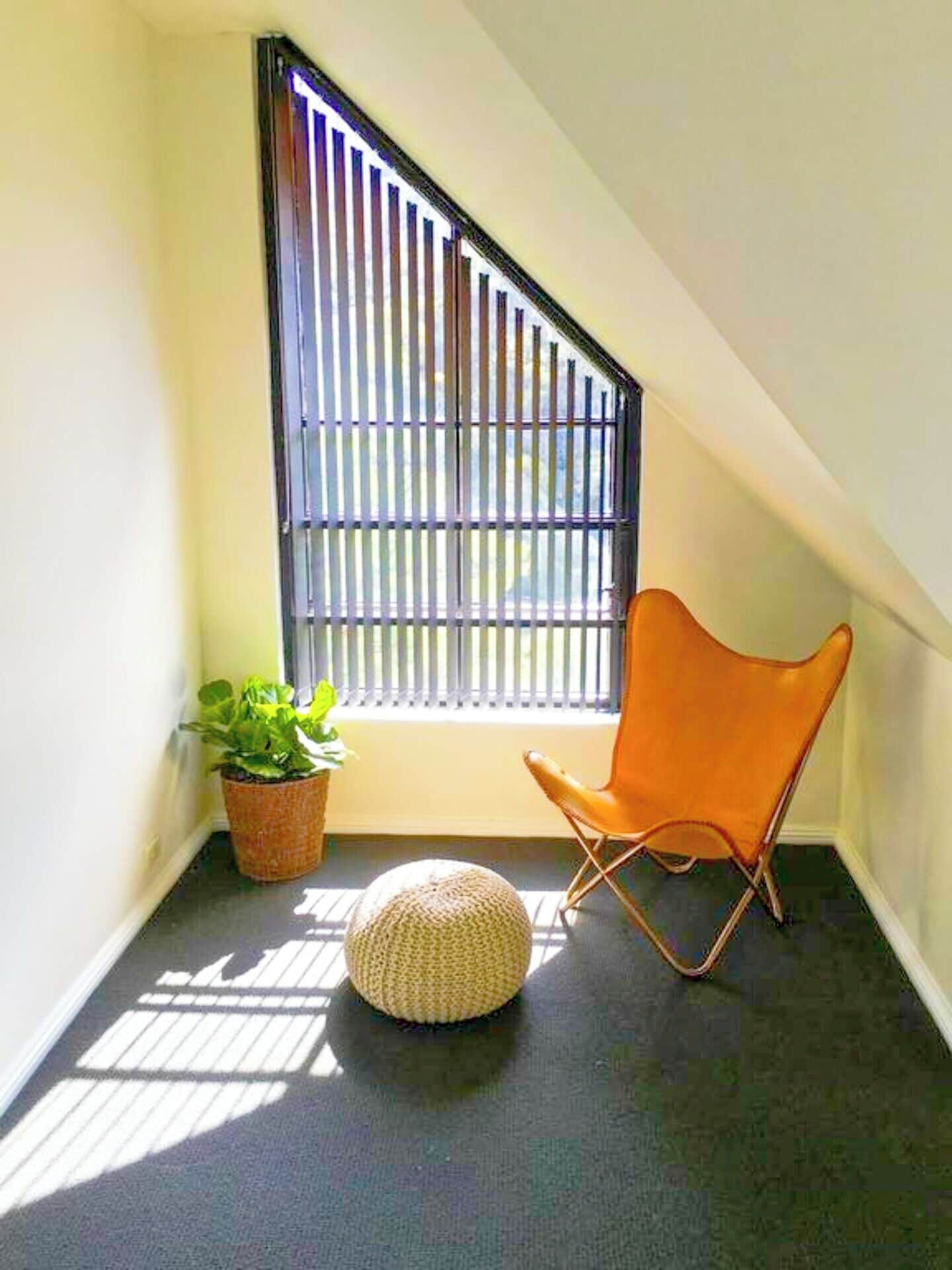Habitació d'angle amb finestra, una planta en test, una cadira i un otomà rodó.