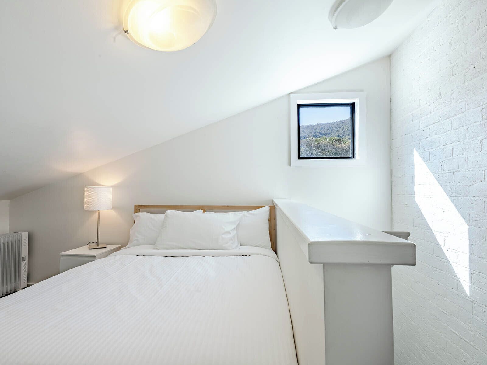 Un dormitorio con techo inclinado, una cama con ropa de cama blanca, una mesita de noche con lámpara y un cuadro enmarcado en la pared.