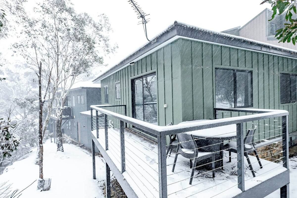 A green wooden cabin with a balcony and a table with chairs, surrounded by snow-covered trees.