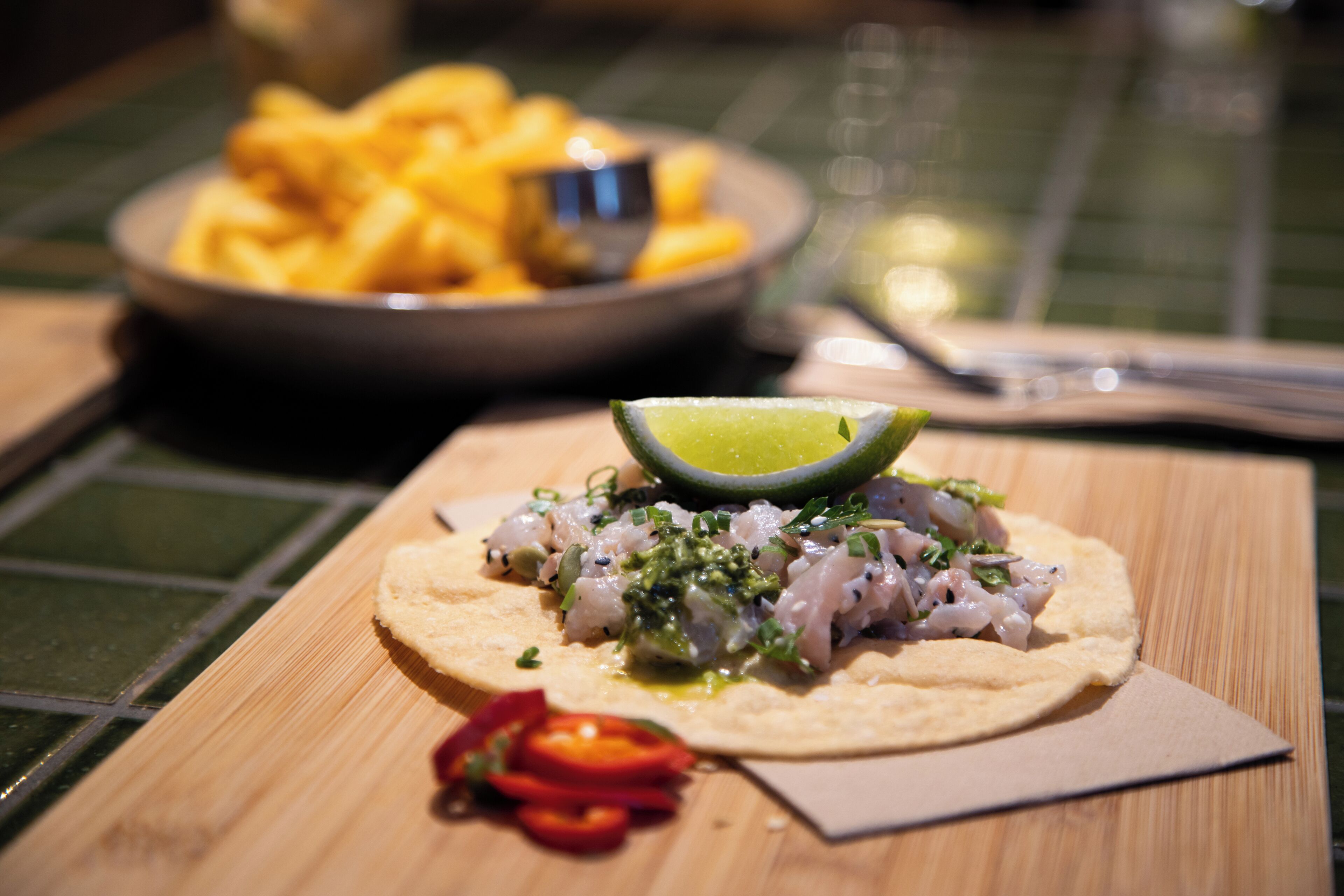A wooden table with a plate of fish tacos, a lime wedge, and a small bowl of fries.