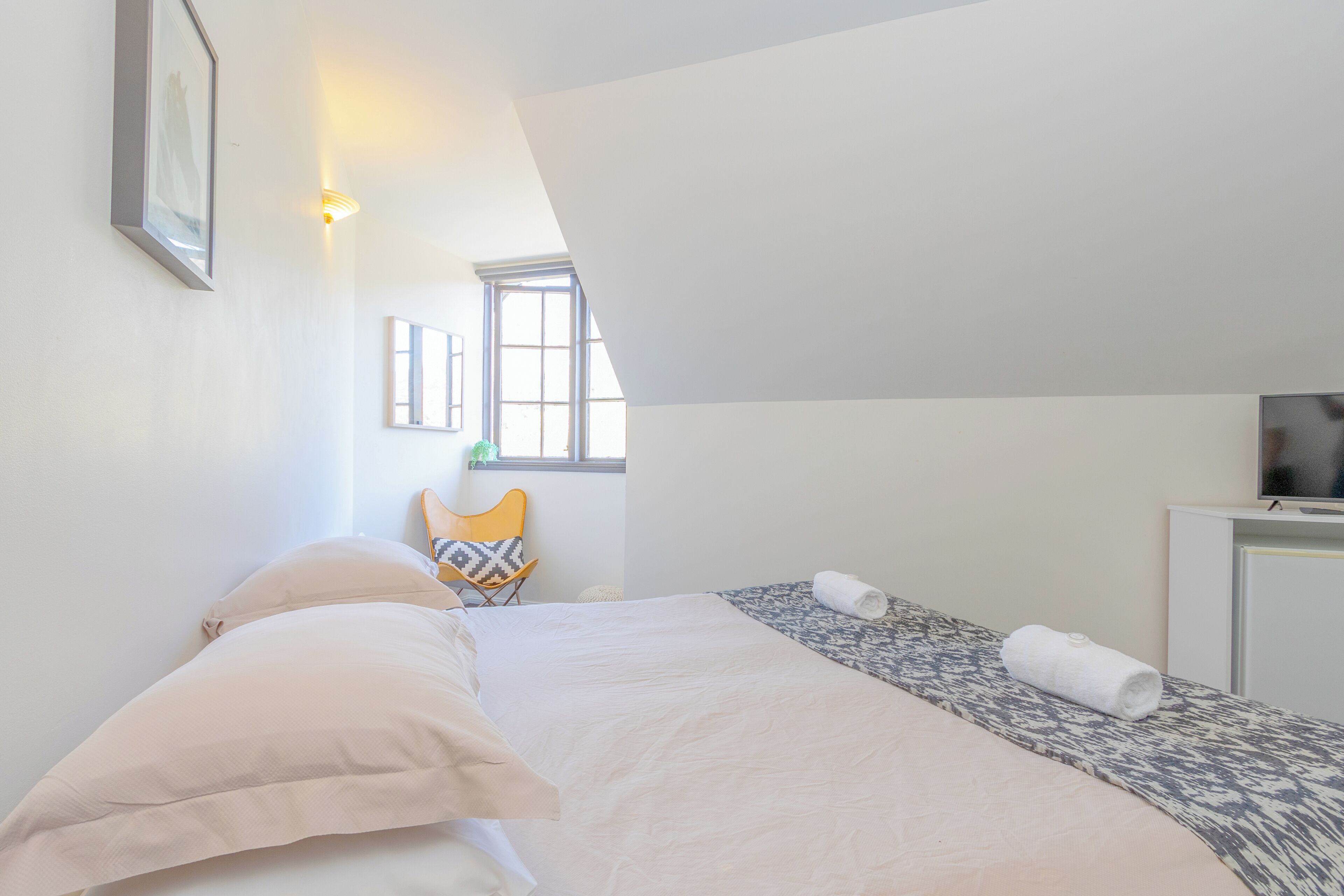 Un dormitorio con cama, una silla, televisor y ventana.