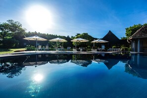 A clear blue pool with sun reflecting on the water, surrounded by lounge chairs and umbrellas, with a thatched-roof building in the background. - Yatule Resort & Spa (Natadola)