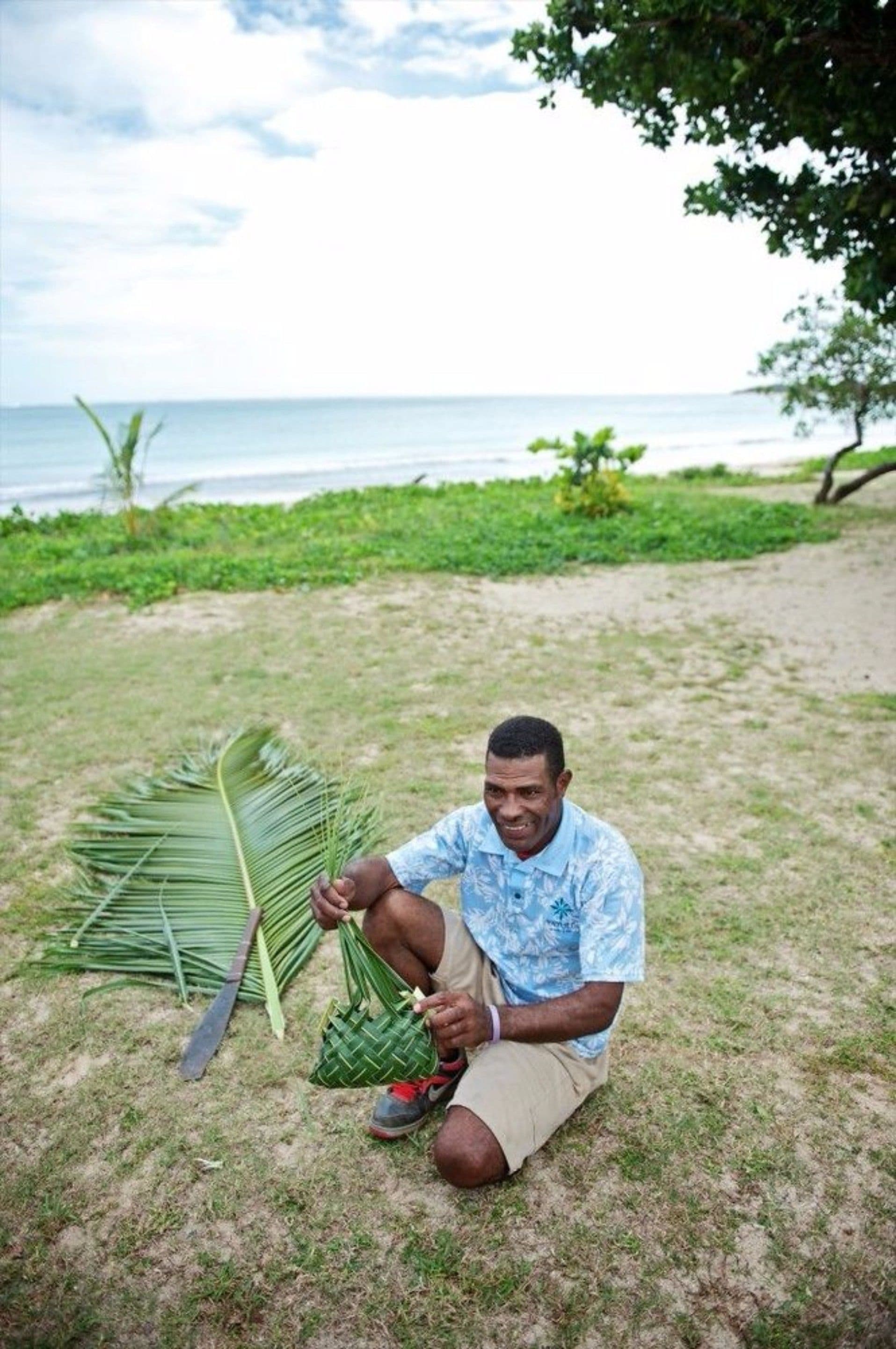 Une personne assise sur l’herbe, près d’une plage, tenant une feuille de palmier et un sac tressé.