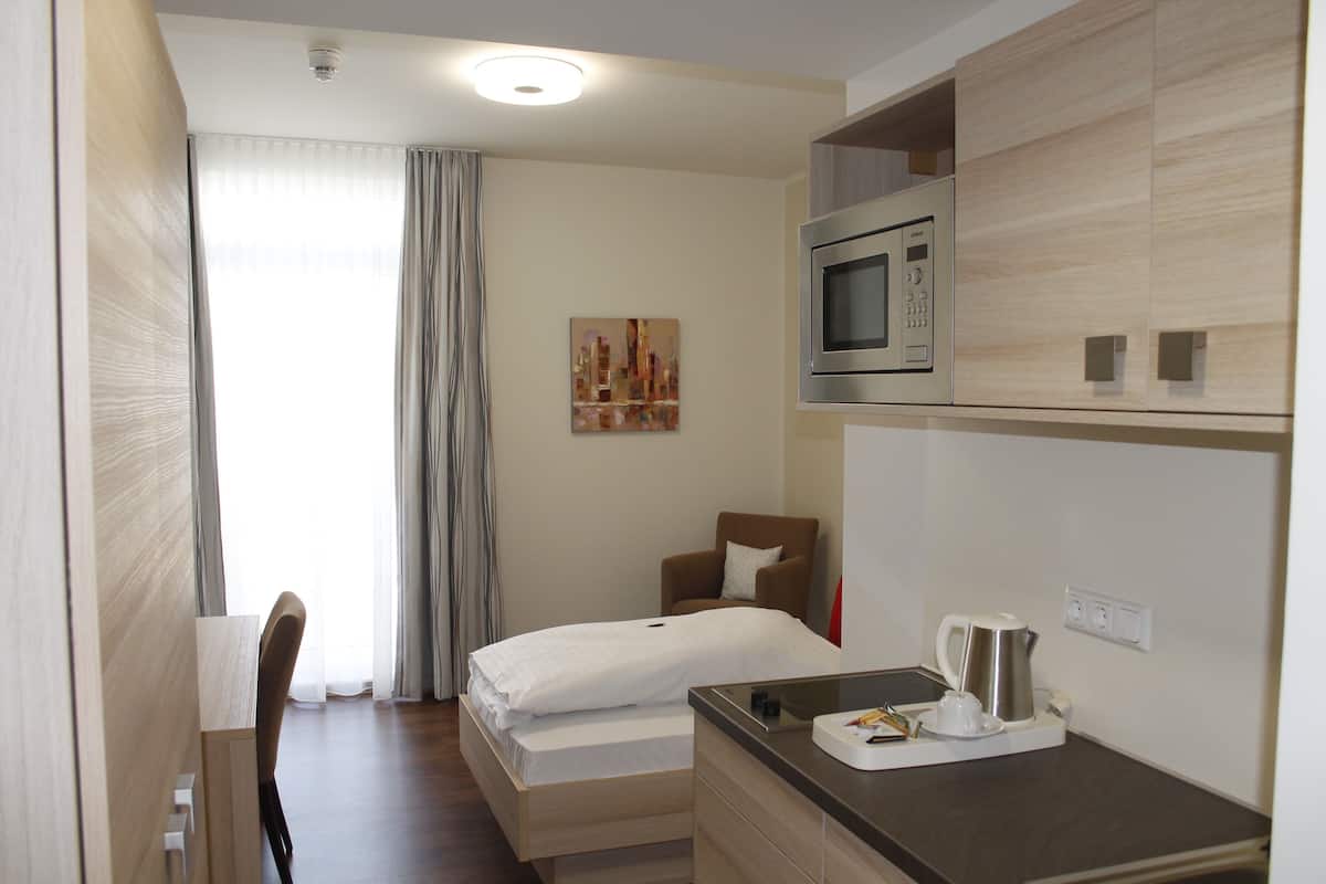 Une chambre d’hôtel avec un lit, une chaise, un bureau avec une bouilloire et des tasses, un four à micro-ondes et un tableau au mur.