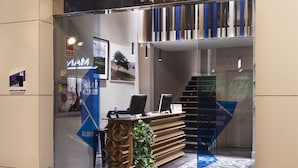 A modern hotel reception with a glass entrance, a reception desk, and a staircase leading to an upper level. - One Shot Colón (Valencia)