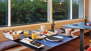 A breakfast setup with a variety of foods and beverages on a table, set against a window with a view of greenery and buildings. - One Shot Colón (Valencia)