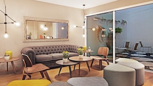 A modern living room with a grey sofa, wooden chairs, and ottomans. There is a large mirror on the wall and a dining area with a table and chairs. - One Shot Colón (Valencia)