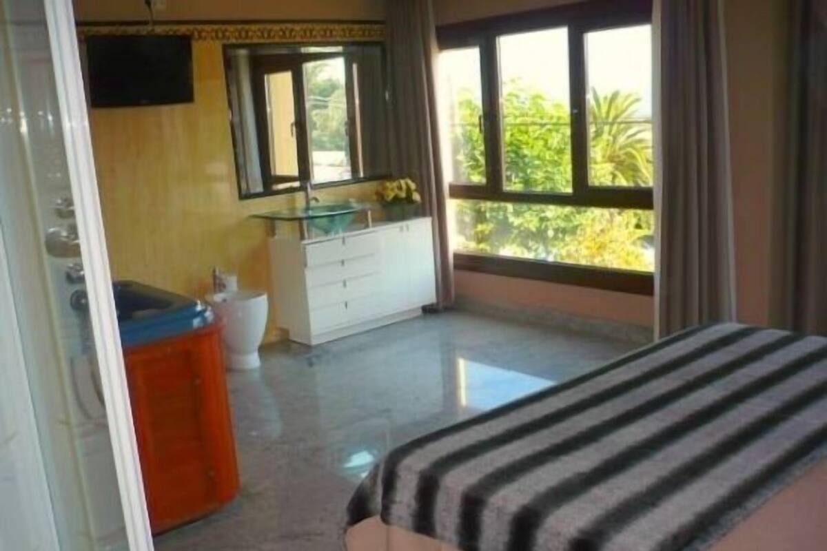 Un dormitorio con cama, un baño con inodoro y lavamanos, y un balcón con vistas a zonas verdes.