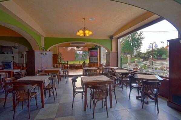 A restaurant with a view of a park, featuring wooden tables and chairs, a large chandelier, and a green and beige color scheme. - Pensión Bos (Bergondo)