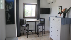 Une petite pièce comprenant un bureau, deux chaises, une commode, une fenêtre avec des rideaux, un téléviseur fixé au mur et un tableau encadré accroché au mur.