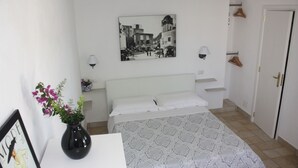 Une chambre à coucher avec un lit, une table de chevet ornée d’un vase de fleurs, une lampe fixée au mur, un tableau encadré et une porte avec un porte-manteau.