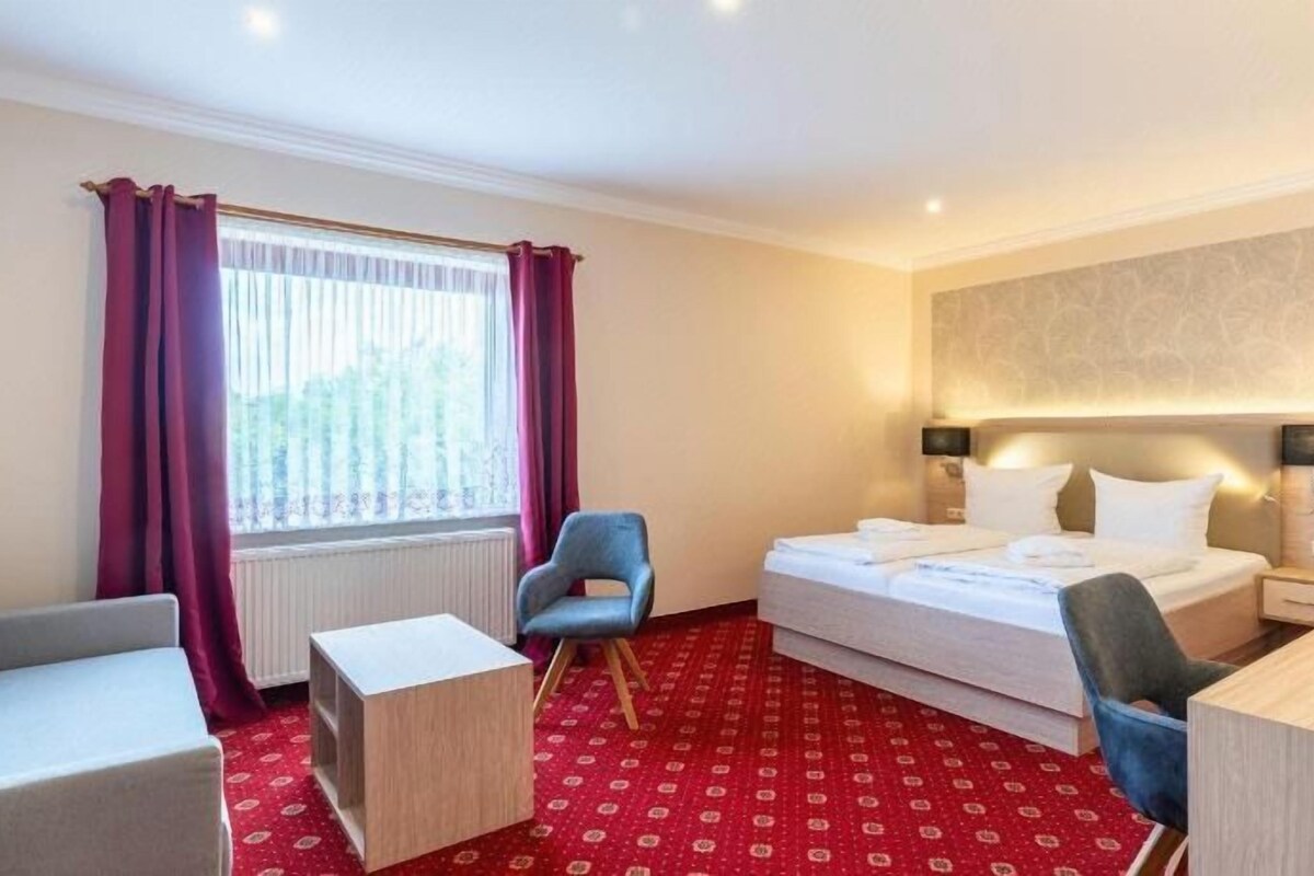 Habitació d'hotel amb un llit, dues cadires, un escriptori, una finestra amb cortines i un radiador.