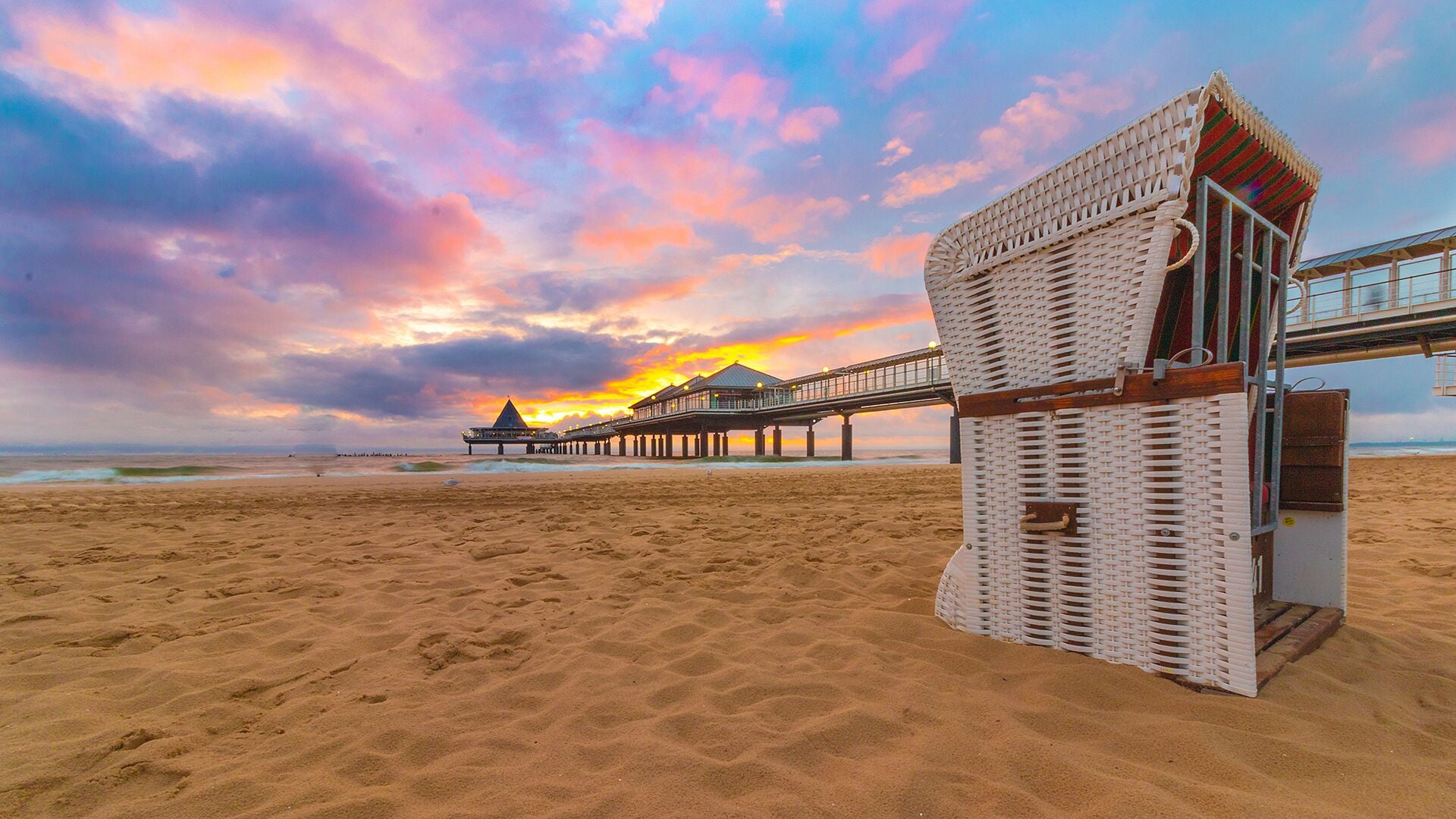 A beach with a wooden pier extending into the ocean, a beach hut, and a sunset in the background.
