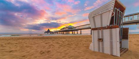 A beach with a wooden pier extending into the ocean, a beach hut, and a sunset in the background.