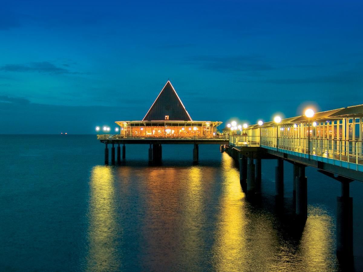 A waterfront hotel with a prominent triangular roof, a pier extending into the water, and a well-lit walkway.