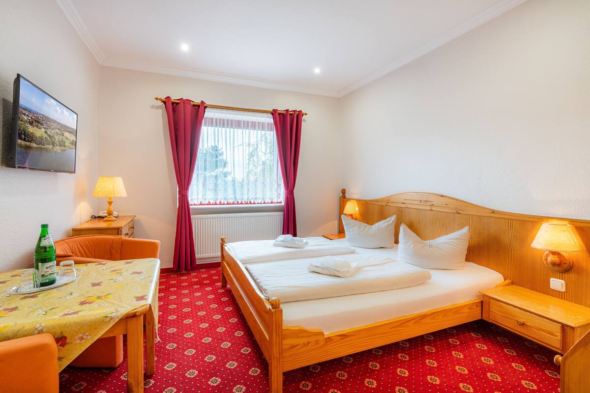 Quarto de hotel com cama de madeira, uma mesa com abajur, uma cadeira e uma janela com cortinas.