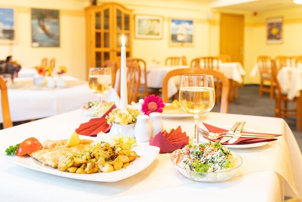 A dining table with plates of food, a glass of white wine, and a candle. - Hotel am Gothensee (Heringsdorf)