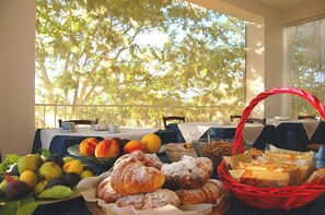 A breakfast table with fruits, pastries, and bread, set against a balcony with trees in the background.