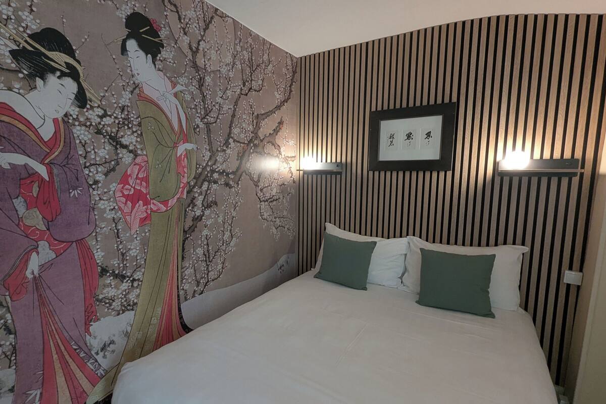 A hotel room with a large mural of cherry blossoms, a bed with white bedding and green pillows, and a wall-mounted light fixture.