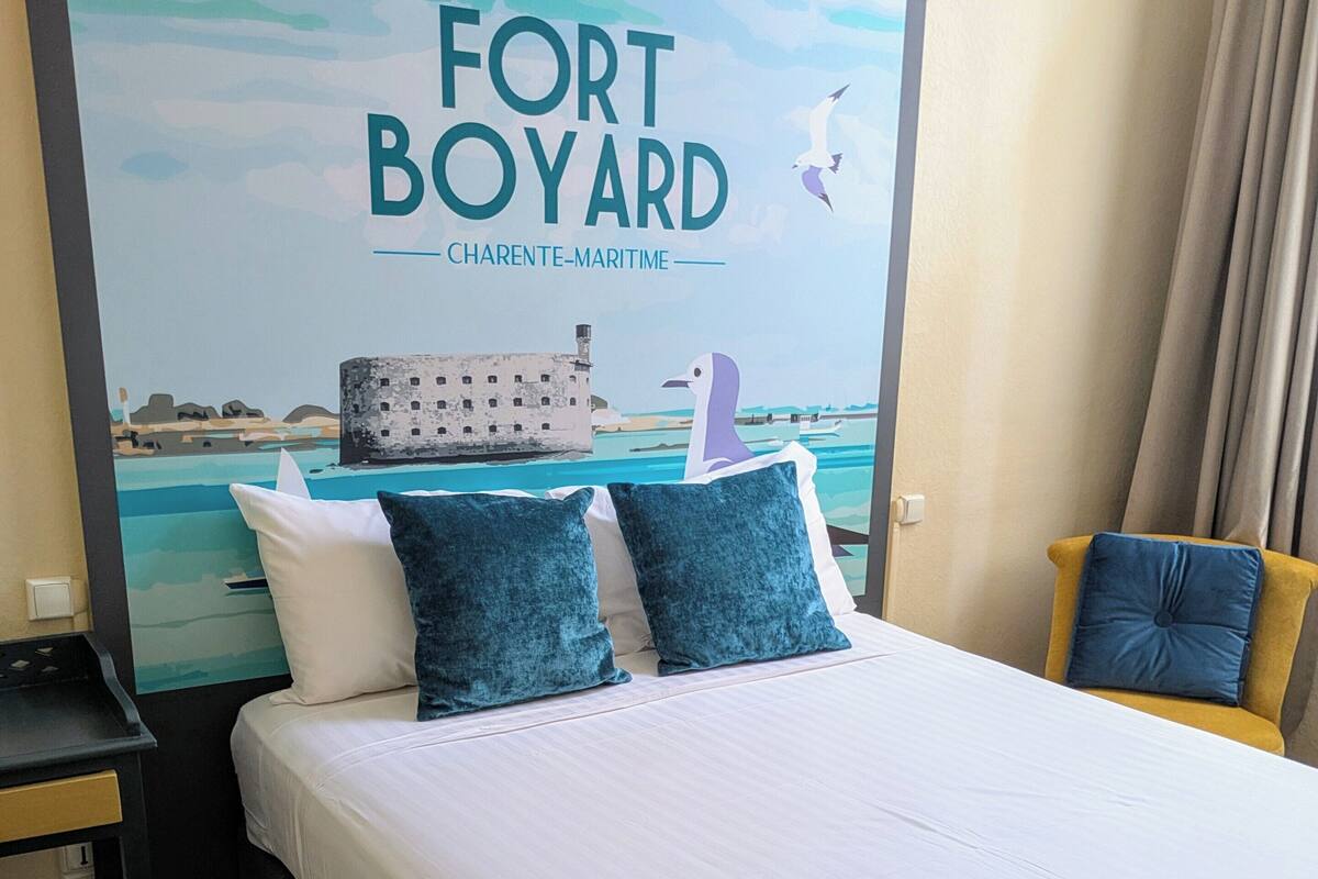 A hotel room with a bed, pillows, a nightstand, and a chair. There is a large poster on the wall with the text "FORT BOYARD CHARENTE-MARITIME".