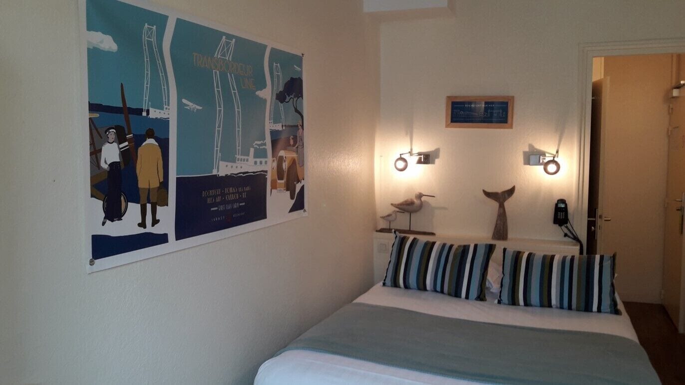 Una habitación de hotel con una cama, un cartel en la pared, un escritorio pequeño con una lámpara y una puerta.