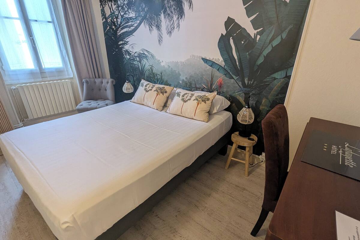 A bedroom with a bed, a chair, a desk, and a large tropical mural.