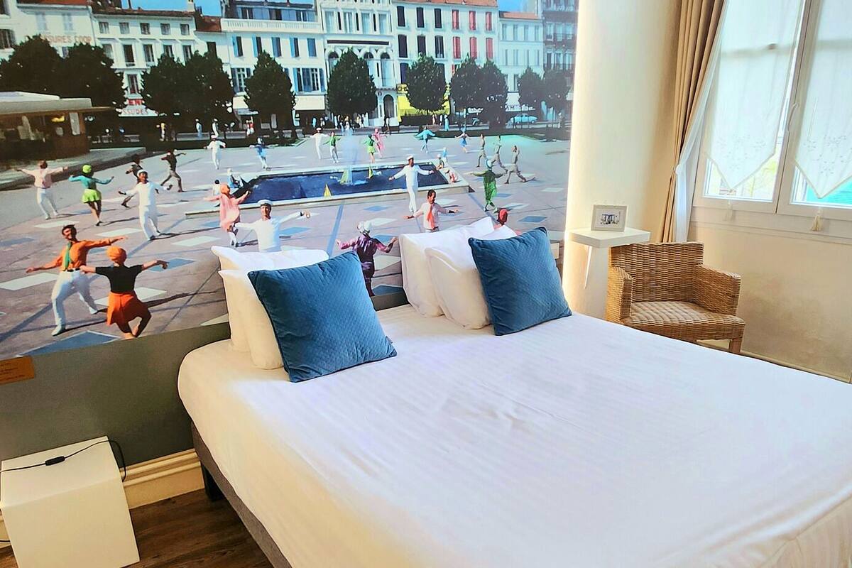 A hotel room with a large bed, two pillows, a wicker chair, and a view of a busy street with people.