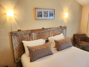 Una cama con cabecera de madera, dos almohadas blancas y dos almohadas a rayas. Encima de la cama hay un cuadro enmarcado y dos lámparas de pared a cada lado.