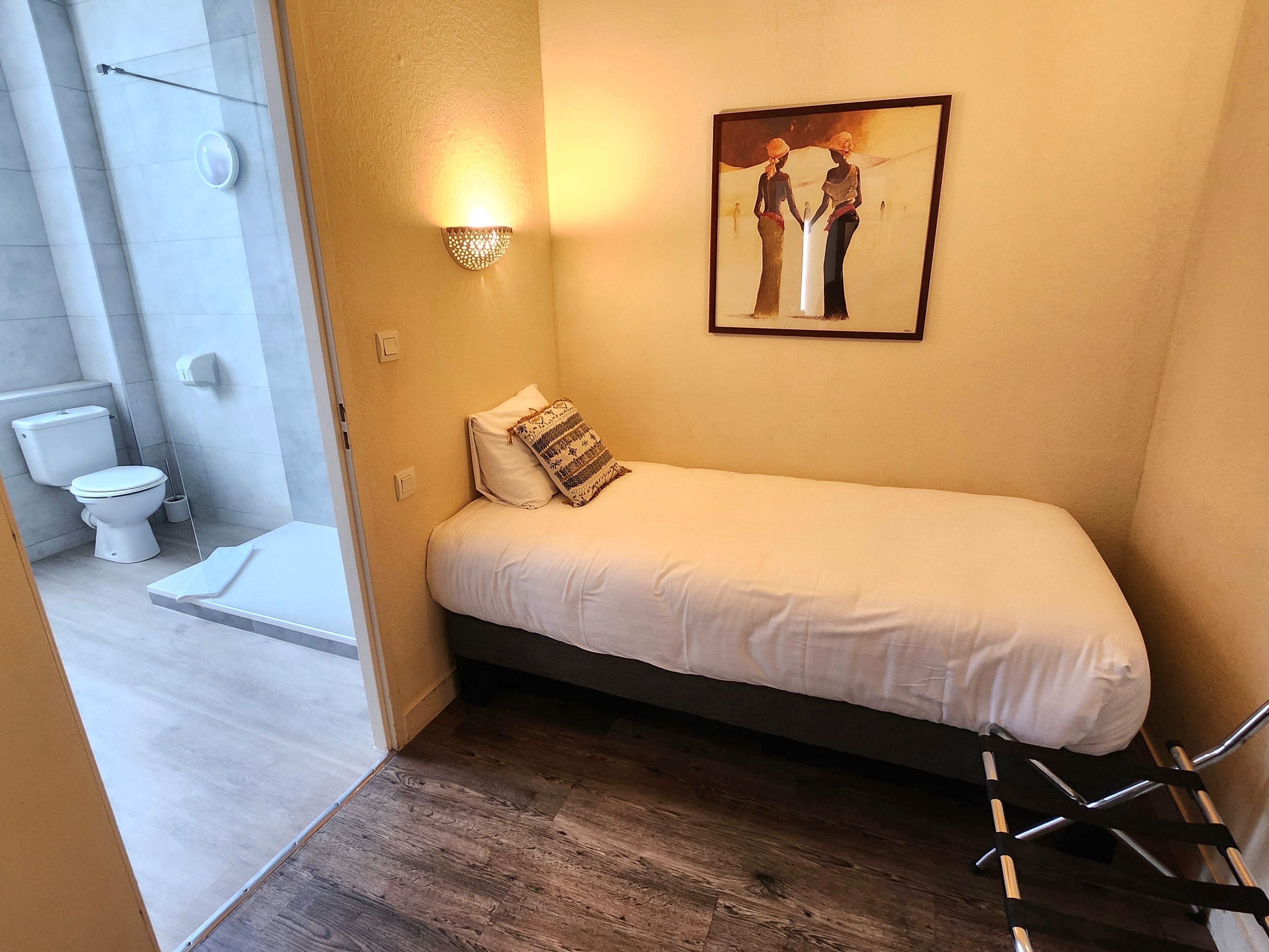 A small, single-bed room with a wooden floor, a white bed, a wall-mounted light, a painting, and a toilet visible in the adjacent bathroom.
