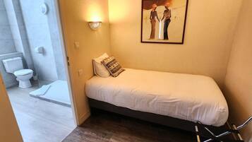 Una habitación pequeña con una cama individual, piso de madera, cama blanca, lámpara en la pared, un cuadro y un baño contiguo con inodoro a la vista.