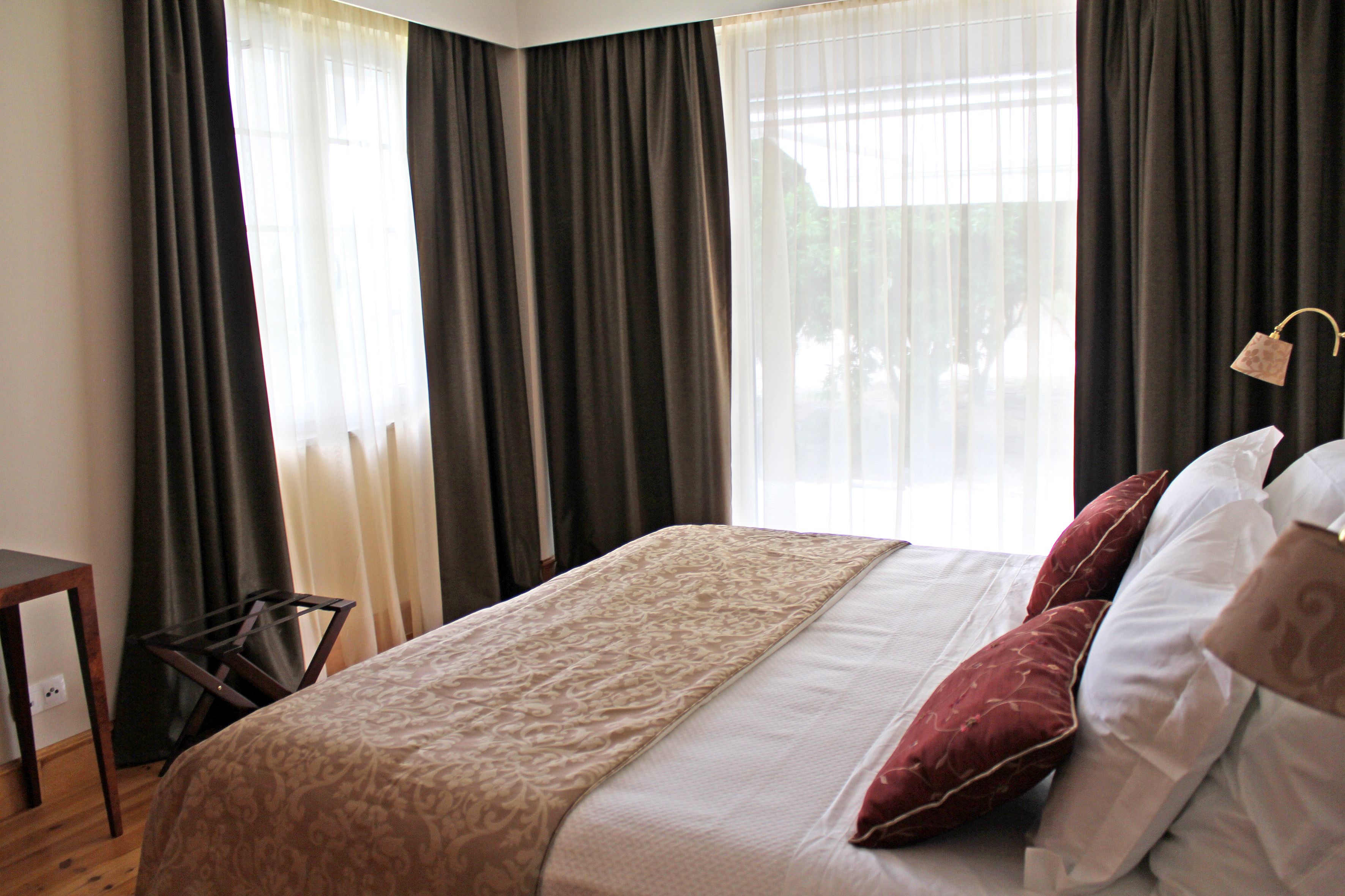 A neatly made bed with a patterned bedspread, a wooden nightstand, and a view of the outdoors through sheer curtains.